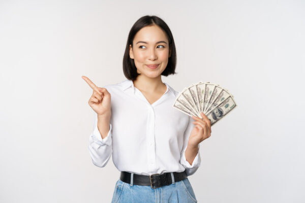 smiling young modern asian woman pointing banner advertisement holding cash money dollars standing white background